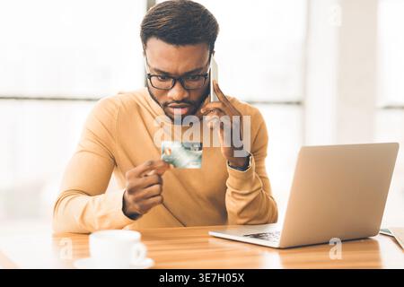 Serious Black man with eyeglasses holding credit card having phone call sitting at table with laptop in office Stock Photo