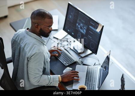 Black male programmer wearing eyeglasses using laptop while sitting at table with computer in office Stock Photo