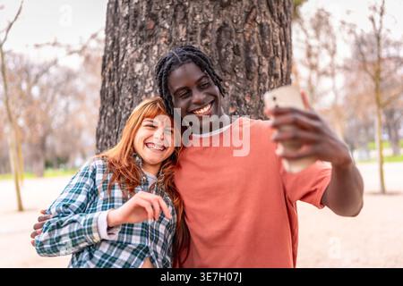 Black man with twists having fun taking selfie on smartphone with caucasian girlfriend while standing under the tree in park during daytime Stock Photo