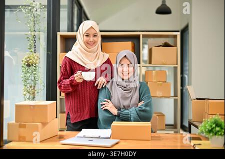 Smiling Muslim female online seller in hijab holding coffee cup standing beside female business pLifestylener sitting with crossed arms at table with car... Stock Photo