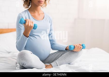 Serious mixed race pregnant woman exercising with small dumbbells sitting crossed legs on bed in bedroom at home Stock Photo