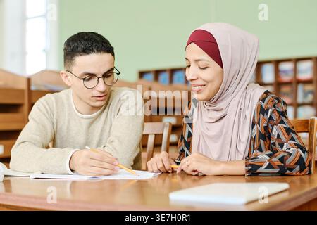 Middle Eastern male and Hispanic Muslim female immigrants studying together while sitting at desk in library Stock Photo