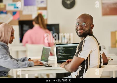Black male programmer looking over shoulder sitting at desk with Mixed race Muslim female colleague working on laptop in office Stock Photo