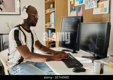 Black man wearing headphones and eyeglasses writing codes on computer while sitting at desk in office Stock Photo