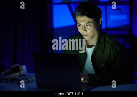 Asian male programmer using laptop while sitting at table working late at night in office Stock Photo
