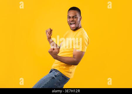 Black man shouting with clenched fists while standing against yellow background in studio Stock Photo