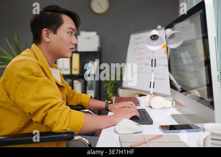 Asian man with disability working on computer sitting in wheelchair in office Stock Photo