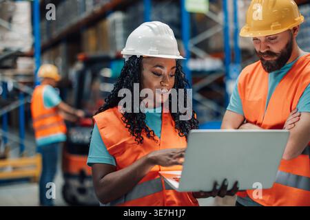 Black female worker wearing hard hat and safety vest showing laptop to colleague standing in warehouse Stock Photo