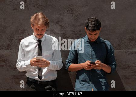 Middle Eastern and caucasian men using smartphones leaning against black wall outdoors on sunny day Stock Photo