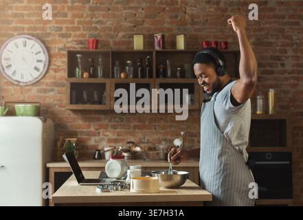 Cheerful Black man in apron listening to music on headphones dancing at table with laptop while baking in kitchen at home Stock Photo