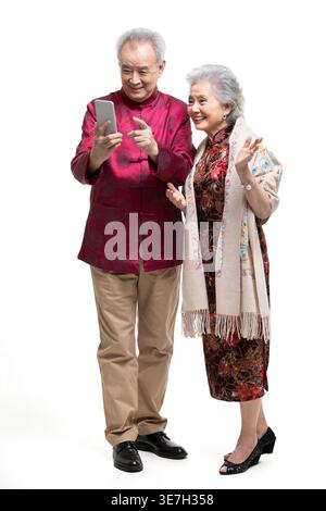 Smiling Chinese senior couple in traditional clothes using smartphone while standing against white background Stock Photo