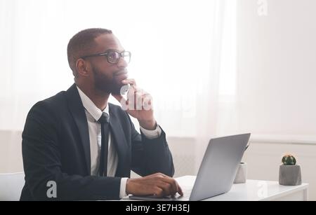 Black businessman wearing eyeglasses touching chin looking away thinking while sitting at desk with laptop in office Stock Photo