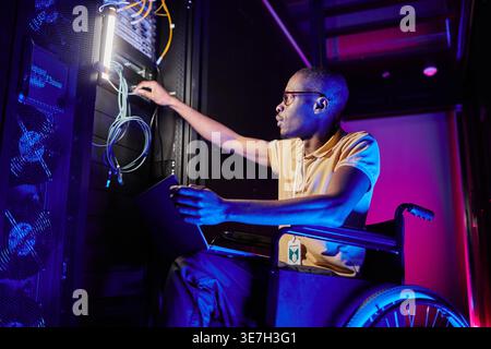 Disabled Black male IT engineer with eyeglasses checking cables sitting on wheelchair with laptop in server room with blue and red neon lights in d... Stock Photo