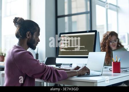 Indian male programmer using laptop near caucasian female colleague sitting at table in office Stock Photo