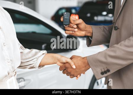 Cropped image of Black client holding his car keys and shaking hands with saleswoamn while standing in car dealerhsip Stock Photo