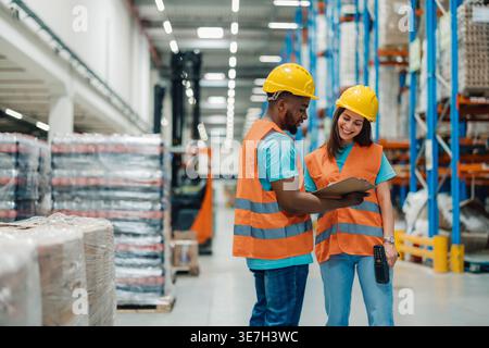Black male worker wearing hard hat and safety vest holding clipboard and managing inventory with caucasian female coworker while standing in warehouse Stock Photo