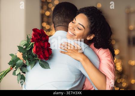Black woman with curly hair holding bouquet of red roses and hugging boyfriend while standing at home Stock Photo