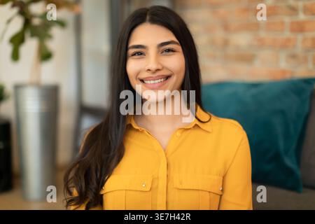 Smiling Middle Eastern woman sitting on floor near sofa in living room at home Stock Photo