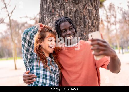 Black man with twists having fun taking selfie on smartphone with caucasian girlfriend while standing under the tree in park during daytime Stock Photo