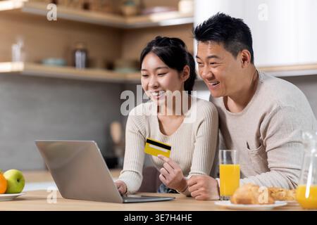 Asian couple using laptop and credit card purchasing goods online while sitting at table with glasses of orange juice and pastries having breakfast... Stock Photo