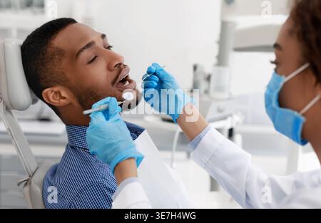 Black male patient getting his teeth checked by female dentist wearing gloves in clinic Stock Photo