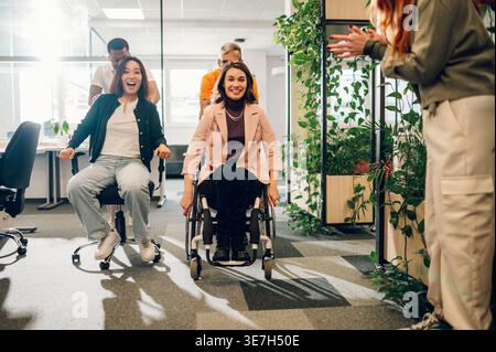 Asian and caucasian female employees having fun playing race sitting in desk chair and wheelchair being pushed by male colleagues in office Stock Photo