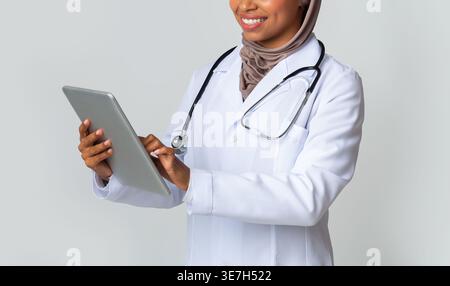 Black Muslim female doctor with stethoscope wearing hijab using tablet while standing against grey background in studio Stock Photo