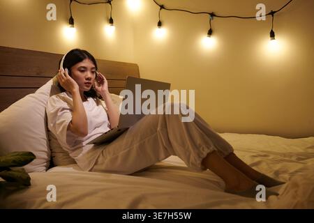 Serious Asian woman wearing headphones watching webinar on laptop sitting on bed at home Stock Photo