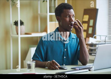Black male office worker using asthma inhaler looking away sitting at table glass of water and document in office during daytime Stock Photo