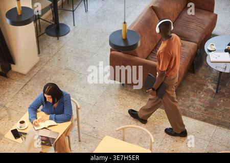 High-angle view of Mixed-race woman writing on pad with laptop and coffee on table while Black man with headphones walking in cafe Stock Photo