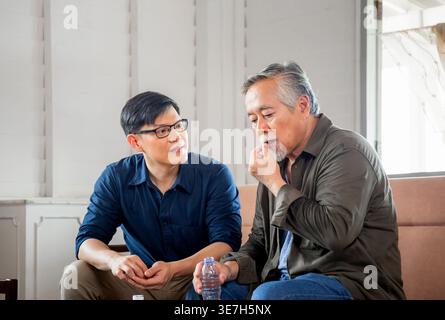 Asian middle-aged man with eyeglasses looking at his senior father drinking medicine sitting beside him on couch at home Stock Photo
