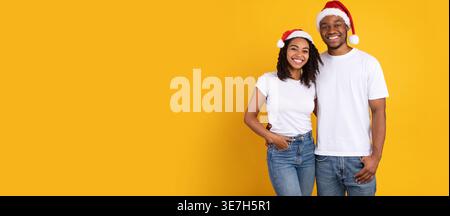 Smiling Black couple wearing Santa hat embracing each other while standing against yellow background in studio Stock Photo