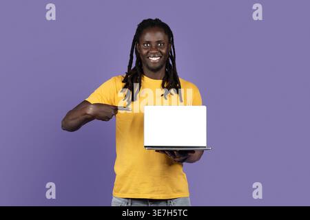 Smiling Black man with dreads wearing yellow shirt pointing at laptop on hand while standing against purple background in studio Stock Photo