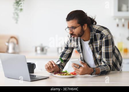 Arab man wearing eyeglasses having snack and looking at laptop while standing at table in kitchen at home Stock Photo