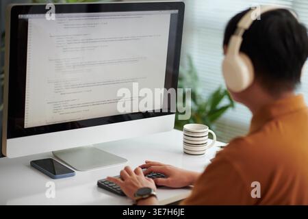 Asian male software developer wearing headphones testing new software on laptop with programming code sitting at desk in office Stock Photo