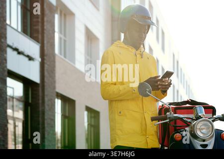 Black delivery man wearing helmet holding smLifestylephone standing near motorcycle with food delivery box on city street during daytime Stock Photo