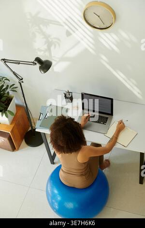 Hispanic woman with curly hair using laptop at desk while sitting on fitness ball at home Stock Photo