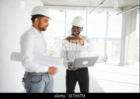Diverse group of male engineers wearing hard hats holding blueprint and laptop discussing project during work on construction site Stock Photo