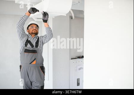 Indian male worker wearing uniform and hard hat preparing ventilation system in an apartment building Stock Photo