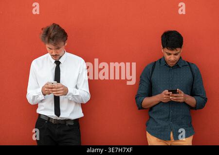 Middle Eastern and caucasian men using smartphones leaning against orange wall outdoors Stock Photo
