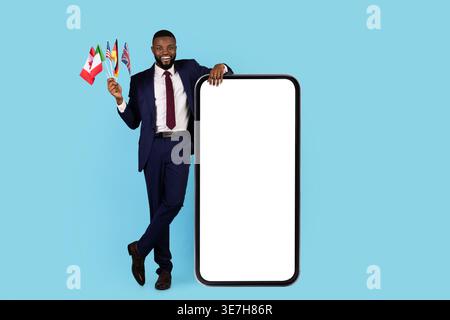 Smiling Black businessman in suit holding different flags leaning on smartphone while standing against blue background in studio Stock Photo