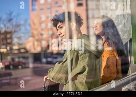 Hispanic couple looking away sitting on bench in bus stop during sunny day Stock Photo