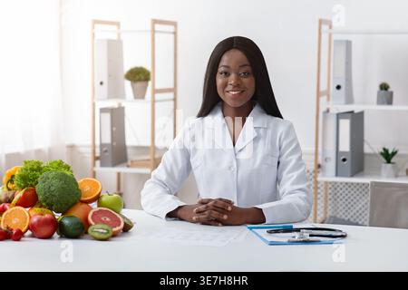 Black female nutritionist sitting with clasped hands at table with clipboard, fruits, and vegetables in clinic Stock Photo