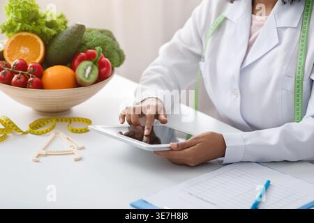 Cropped image of Black female dietitian with tape measure around her neck using tablet while sitting at table with bowl of fruits and vegetables in... Stock Photo