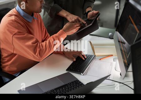 Mixed-race female programmer working on tablet with caucasian male colleague while sitting at table with laptop in office Stock Photo