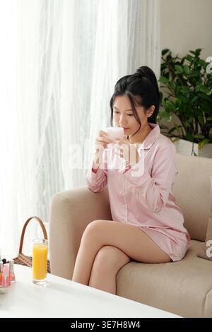 Asian woman in silk pink pajamas smelling jar of moisturizing body cream while sitting on sofa at home Stock Photo