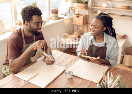 Black male artist wearing apron and eyeglasses holding brush and palette explaining pottery details with female student sitting at table in workshop Stock Photo
