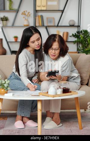 Middle-aged Asian mother and daughter computing their household bills using calculator while sitting on sofa near table in living room at home Stock Photo