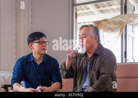 Asian middle-aged man looking at his senior father drinking water sitting beside him on couch at home Stock Photo