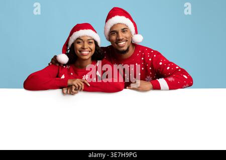 Smiling Black couple wearing Santa hats and sweaters leaning on white board against blue background in studio Stock Photo